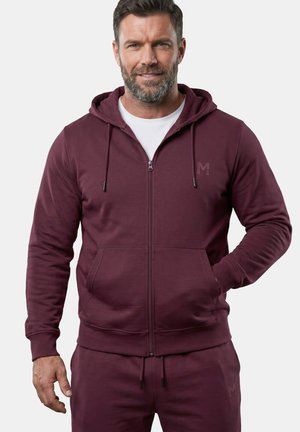 Man met baard die een donkerrode hoodie met rits en bijpassende joggingbroek draagt, staand met één hand in de zak tegen een effen achtergrond.
