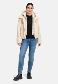 Veste extérieure beige et duveteuse avec capuche, superposée à un gilet crème. Portée avec un haut noir, un jean bleu et des bottines à talons noires.