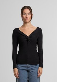 Top negru cu mâneci lungi, decolteu în V și detaliu răsucit în centru. Textură ribanată, croială mulată, asortat cu blugi bleu deschis.