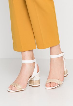 Voeten met witte sandalen met blokhak en enkelbandje, voorzien van geweven beige details, gecombineerd met mosterdgele cropped broek.