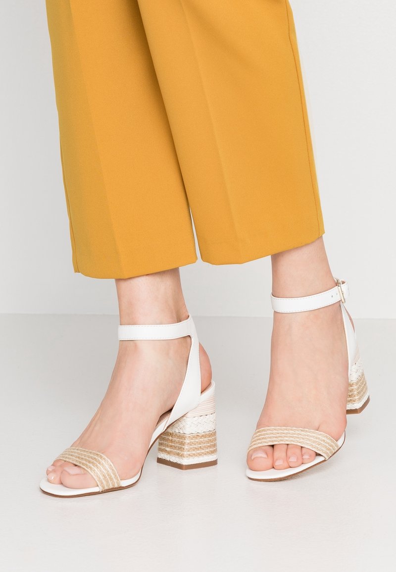 Pieds portant des sandales à talons blocs blanches à bride cheville avec des détails en beige tissé, associées à un pantalon court jaune moutarde.