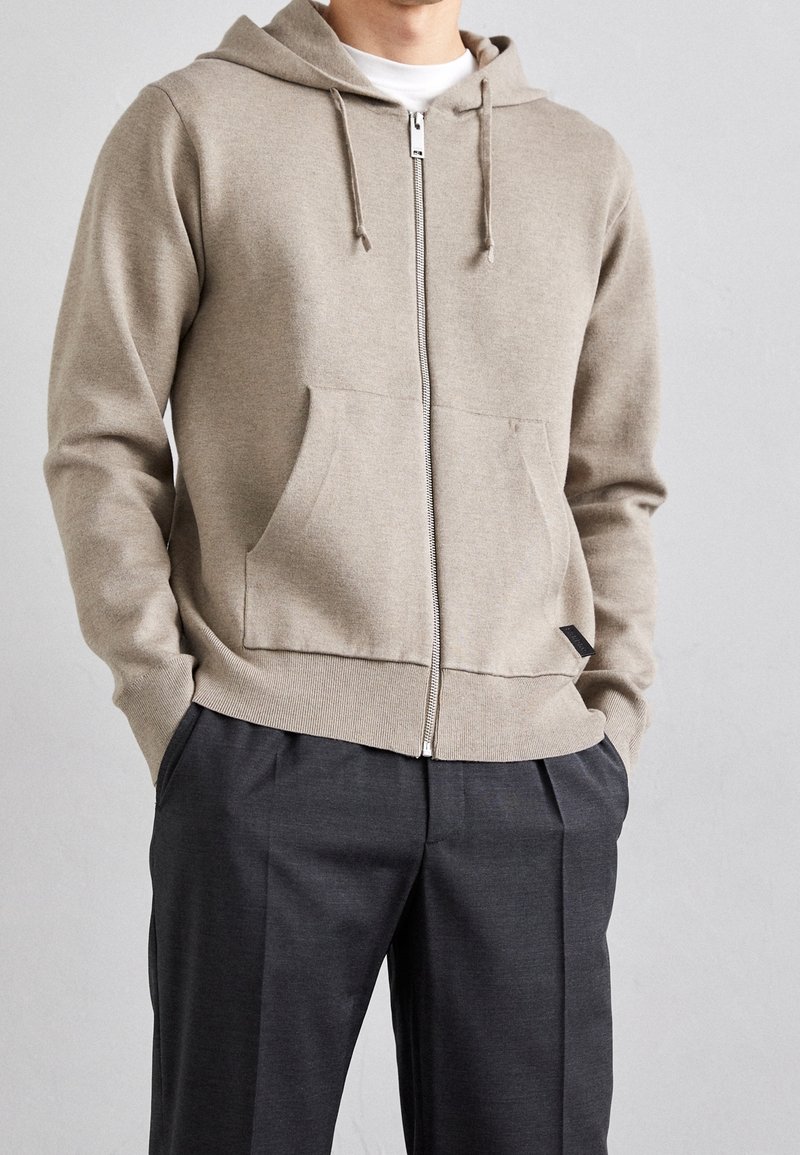 Hellbeige Zip-Up-Hoodie aus weichem Material, mit einer vorderen Tasche, Kapuze mit Kordelzug und Rippbündchen, kombiniert mit dunkelgrauen Hosen.