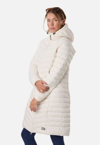 Jeune femme aux cheveux châtain clair portant un long manteau matelassé blanc avec capuche, des leggings bleu marine et des lunettes de soleil sur la tête.