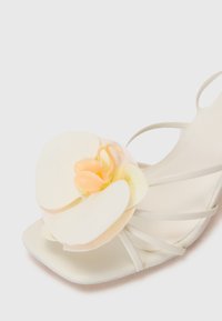 Sandalino bianco con cinturini, caratterizzato da un'ampia decorazione di orchidea crema e gialla pallida. Texture liscia con un design a punta quadrata.