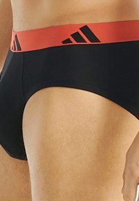 Mustat alushousut, joissa on punainen elastinen vyötärönauha, ja mustia raidallisia yksityiskohtia sekä Adidas-logo. Su гладка pinta, tiukka muoto.