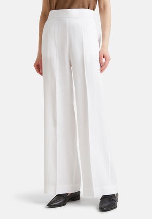 Kobieta ubrana w białe spodnie z wysokim stanem i szerokimi nogawkami, do tego czarne loafersy i brązowy top, stoi na tle jednolitego tła.