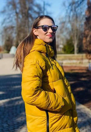 Žlutá péřovka s vysokým límcem a zipem, s prošívanou texturou. Černé sluneční brýle a dlouhé, rovné vlasy. Venkovní prostředí.