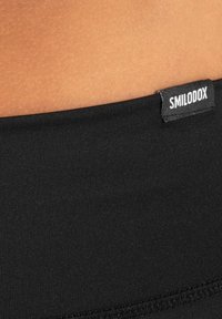 Close-up of black fabric garment with SMILODOX label sewn near the edge against light brown skin background.