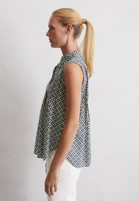 Ärmellose Bluse mit Kragen, die ein blaues geometrisches Muster mit gelben Akzenten zeigt. Aus leichtem Stoff, leicht lockerer Schnitt.