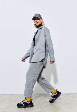 Cămașă din bumbac gri cu butoni supradimensionată și pantaloni asortați dintr-un material texturat. Adidași negri cu accente galbene. Șapcă gri cu un model în rețea.