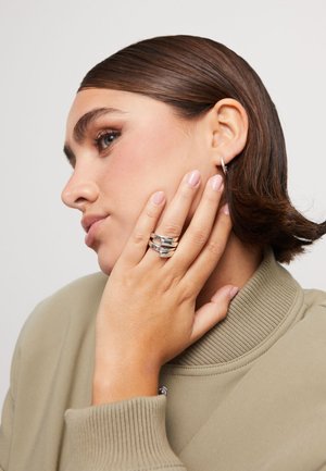 Joven con cabello corto y castaño que lleva un pendiente de aro plateado y tres anillos anchos de plata, tocándose la cara con la mano.