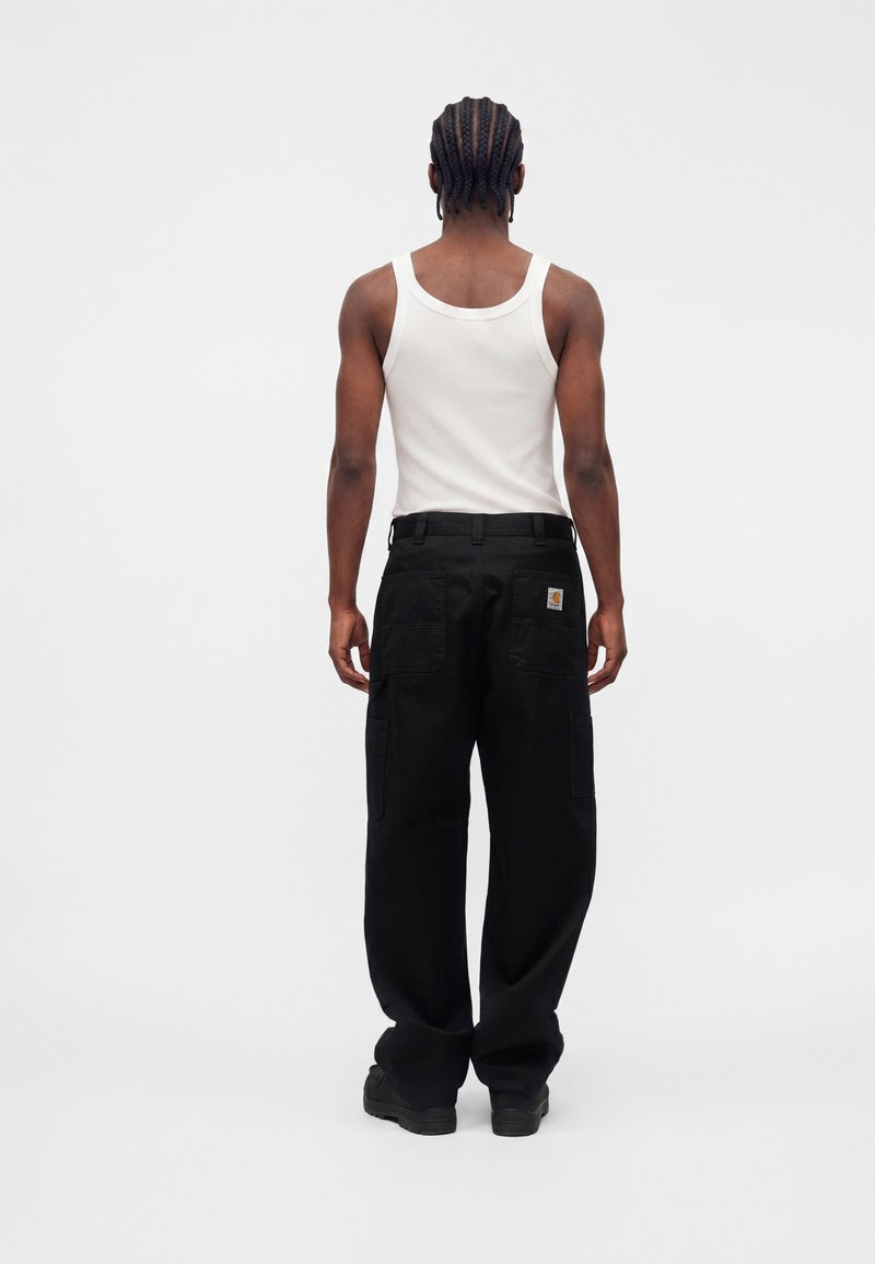 Person with braided hair wearing a white tank top, black cargo pants, and black shoes, standing facing away against a plain background.