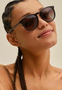 Occhiali da sole neri con lenti rotonde, aste sottili e design minimalista; indossati da una persona con capelli intrecciati, su uno sfondo neutro.
