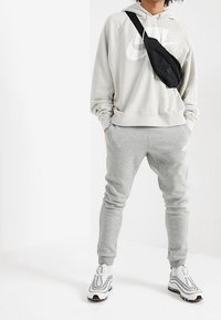 Grå huvtröja med stor vit logotyp, matchande grå joggers, svart slingväska och vita sneakers med texturerade detaljer och grå accenter.