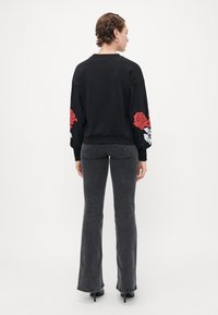 Kvinne med kort hår som har på seg en svart hettegenser med røde og hvite roseprint på ermene, mørke grå bukser og svarte hæler, står.