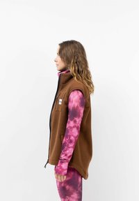 Bruine fleece vest met hoge kraag en ritssluiting aan de voorkant, gedragen over een roze tie-dye langemouwen top. Bevat zijzakken en een logo patch.