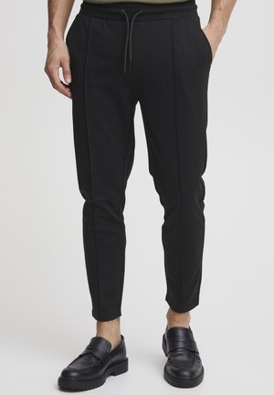 Mann trägt schmale schwarze Jogginghosen mit elastischem Bund und Kordelzug, kombiniert mit schwarzen Leder-Loafers, steht mit den Händen in den Taschen.