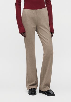 Personne portant un pantalon beige droit, des mocassins noirs et un haut rouge à manches longues avec des manches allongées sur un fond uni.