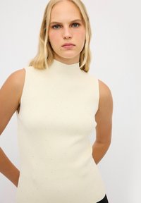Mouwloze, geribbelde top in lichtcrème met een hoge hals. Bevat kleine, verspreide zilveren stippen voor textuur en details.