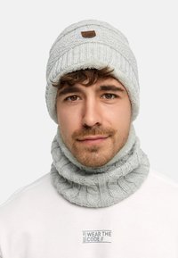 Light gray knit beanie with a patterned design and a brown leather patch, paired with a matching ribbed neck warmer. Soft texture.