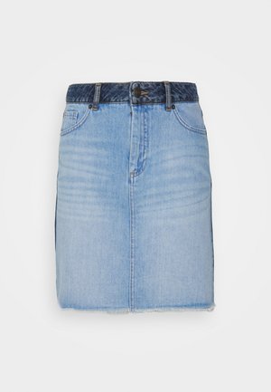 Nainen, jolla on päällään lyhythihainen kiiltävä kultainen paita, vaaleansininen denimhame ja mustat paksupohjaiset nauhalliset kengät, seisoo valkoista taustaa vasten.