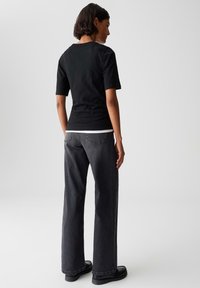 Donna in piedi di spalle, indossa una maglietta nera a maniche corte, jeans larghi grigio scuro e scarpe nere, su uno sfondo grigio uniforme.