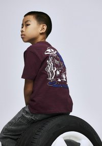 Bordowa bawełniana koszulka z białym i niebieskim nadrukiem przedstawiającym kaktusy, UFO oraz tekst na plecach, w połączeniu z szarymi dżinsami, siedząca na oponie.