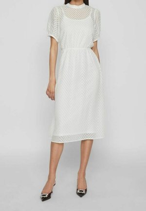Femme portant une robe midi blanche semi-transparente avec des manches ballon et un motif chevron, associée à des talons pointus noirs avec des ornements décoratifs.