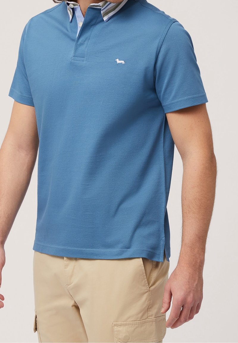 Polo bleu à manches courtes avec une patte de boutonnage à deux boutons. Il présente un col rayé et un petit logo blanc sur la poitrine.
