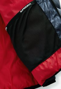 Chaqueta roja y negra con forro de malla, detalles de material brillante y un parche de logo prominente en la manga.