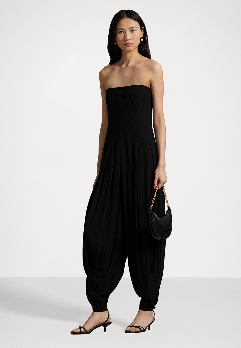 Donna in tuta nera senza spalline con pantaloni plissettati e larghi, sandali neri con tacco, orecchini di perle e piccola borsa nera con tracolla dorata.