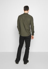 Camisa de manga larga verde oscura con puños abotonados y un yugo en la parte posterior, combinada con pantalones negros. El conjunto tiene un ajuste relajado y un diseño minimalista.