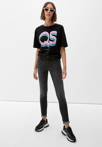 Camiseta negra con un gráfico colorido de "QS", combinada con jeans ajustados gris oscuro y zapatillas negras con detalles en blanco. Gafas de sol blancas en forma de ojo de gato.