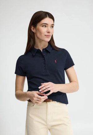 Vrouw draagt een marineblauw poloshirt met korte mouwen en een roze logo, beige broek en een bruine riem, staand tegen een effen witte achtergrond.