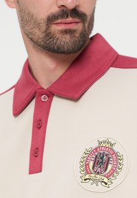 Άνδρας που φοράει μπεζ πόλο πουκάμισο με μπορντό γιακά και κουμπιά, με έμβλημα του Varsity Athletic Society στο στήθος.