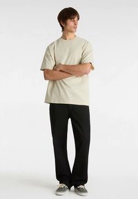 Beige bomuld t-shirt med løs pasform og korte ærmer, kombineret med sorte bukser. Personen bærer mønstrede sneakers.