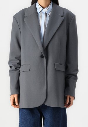 Personne portant un blazer gris à un bouton sur une chemise à col rayée bleue et blanche, et un jean bleu foncé, debout devant un fond blanc.