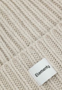 Zbliżenie kremowej, prążkowanej dzianiny z białą prostokątną metką oznaczoną napisem "Elementy", przyszytą w dolnym prawym rogu.