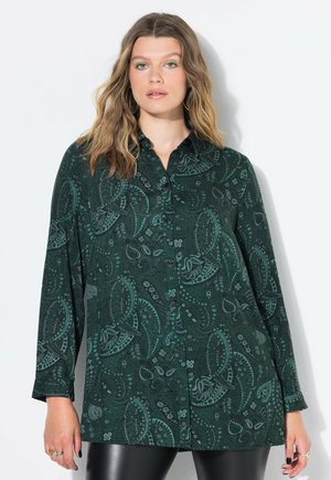 Donna che indossa una camicia button-up a paisley verde scuro, pantaloni di pelle neri, con capelli mossi e sciolti, orecchini a cerchio, eyeliner verde e anelli d'argento.