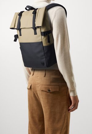 Personne portant un sac à dos beige et noir à fermeture roulante avec des bretelles noires, associé à un pantalon beige et un haut à manches longues beige.