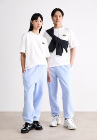Două persoane poartă tricouri albe cu accente verzi ale logo-ului, asortate cu pantaloni de trening bleu deschis. Una dintre ele are un hanorac bleumarin aruncat pe umeri.