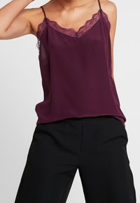 Burgundyröd satin camisole med spetsdetaljer, som har reglerbara spagettistroppar och en avslappnad passform, i kombination med svarta högmidjade byxor.