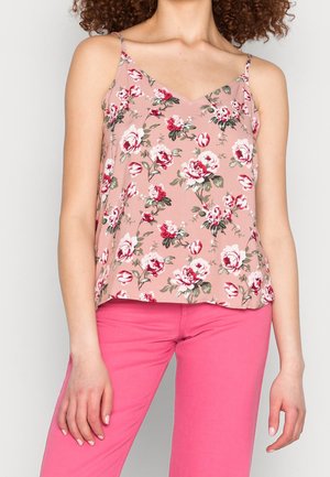 Kobieta w różowej kwiecistej bluzce na ramiączkach i jaskraworóżowych spodniach, stojąca na jednolitym tle.