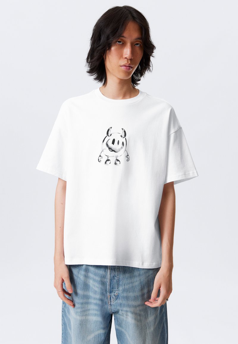 Tricou alb cu mânecă scurtă, din bumbac, având un grafic cu un personaj de desene animate de culoare neagră pe față. Modelul poartă blugi din denim albastru.