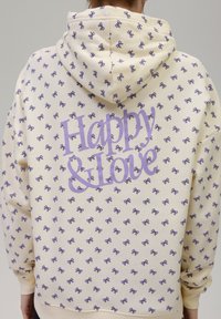 Sudadera crema con un estampado de lazo púrpura en toda su superficie. Presenta el texto "Feliz y Amor" en púrpura en la parte de atrás, con capucha ajustable.