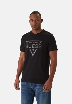 Mężczyzna stojący w czarnej koszulce Guess z logo oraz niebieskich dżinsach na jasnym, jednolitym tle.