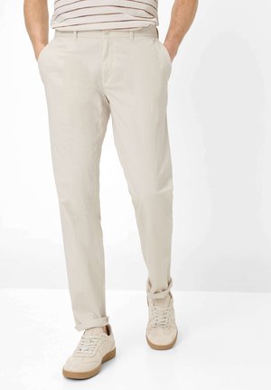Mann trägt beige Chinos, cremefarbene Sneakers und ein gestreiftes Hemd, das in die Hose gesteckt ist, steht mit den Händen in den Taschen vor weißem Hintergrund.