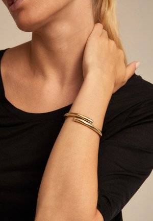 Bracelet en or avec un design ondulé et poli, ajusté sur un poignet, sur un fond neutre et porté avec un haut noir.