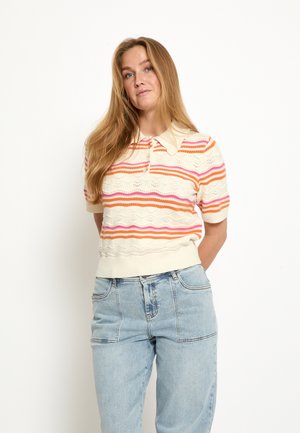 Pitkähiuksinen vaalea nainen, joka käyttää kermanväristä neulepuseroa, jossa on oransseja ja vaaleanpunaisia raitoja, sekä vaaleansinisiä korkeavyötäröisiä farkkuja, seisoo valkoista taustaa vasten.