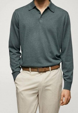 Mand iført en afdæmpet grøn langærmet polo skjorte stoppet ned i beige bukser med et brunt bælte, stående med den ene hånd i lommen.